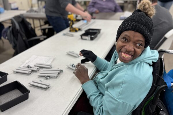 A black woman in a wheelchair wearing a winter hat sits at a table smiling, as she works to assemble pieces of metal track.