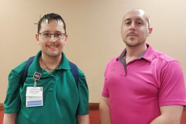 Matt is on the left, he has short brown hair and glasses and is smiling. He wears his work badge on his green polo shirt. His job coach, Casey, is on the right, with a smooth bald head, and wearing a pink polo shirt.